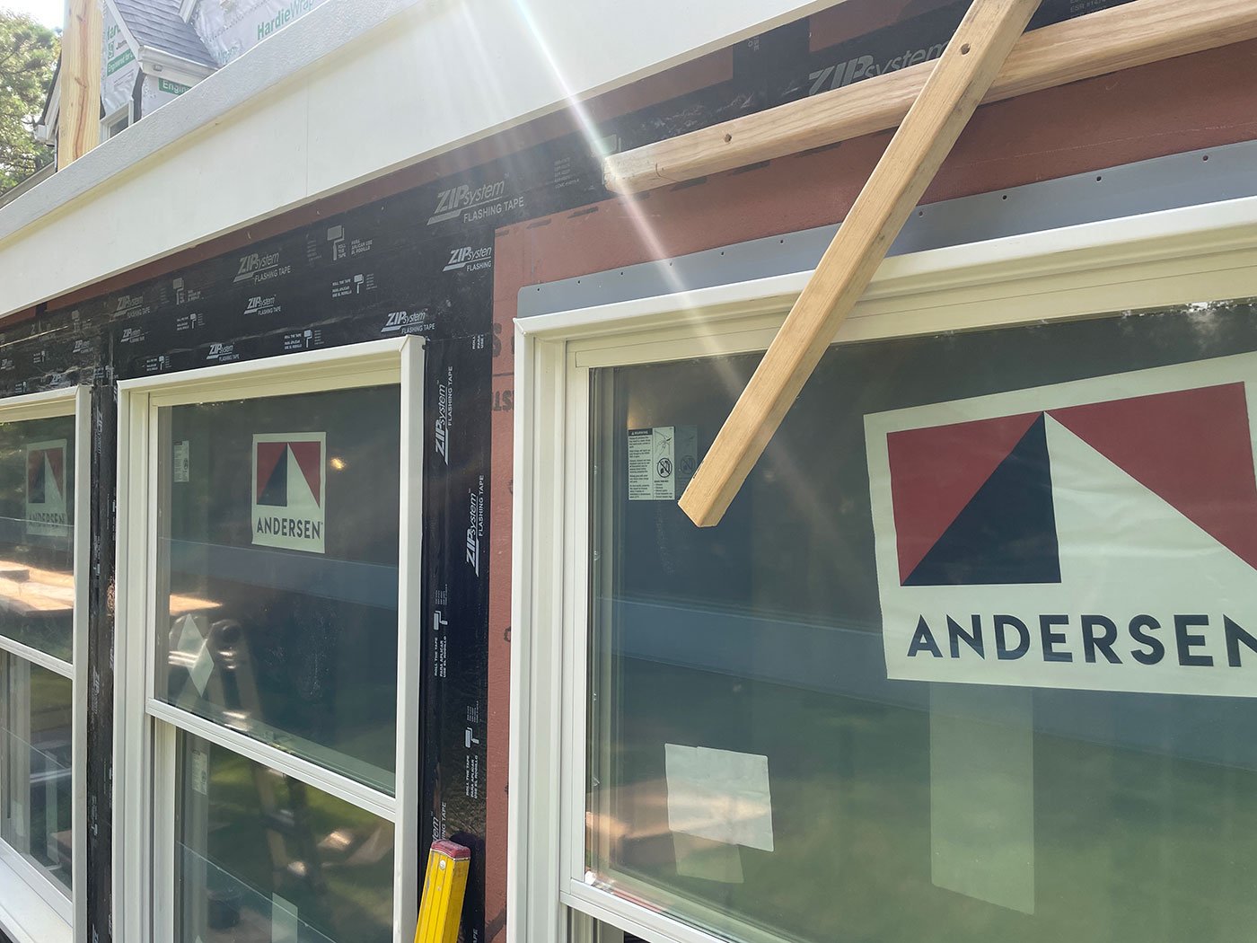 window that has been installed on a wall that has signs of incomplete exterior work. The wall framing and surrounding materials are exposed, and a board with the "Andersen" brand is visible in the window. A diagonal wooden beam is leaning against the wall, suggesting ongoing repairs or construction.