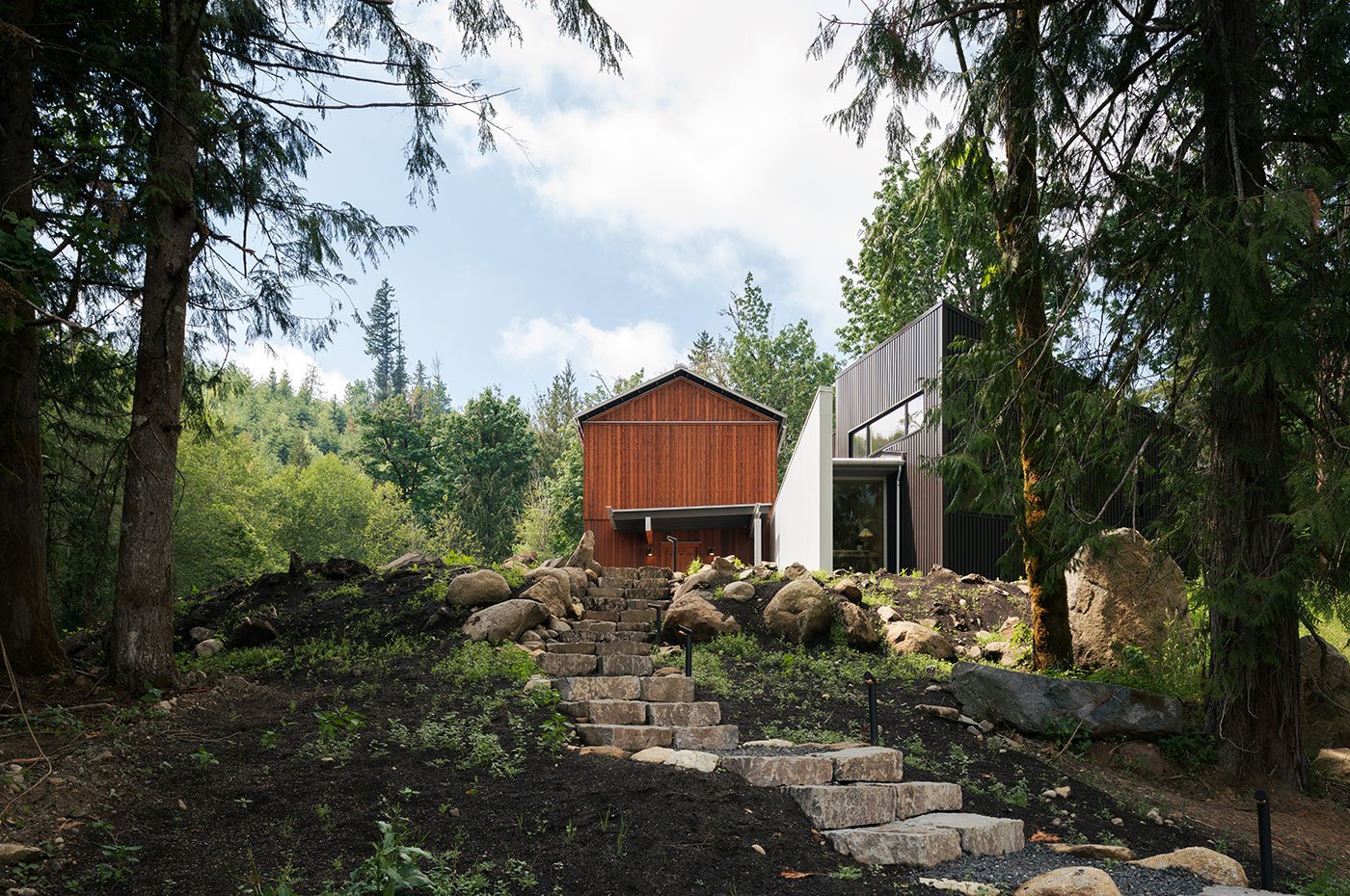 A stone stairway leading to a home made of two distinct volumes, one clad in cedar and the other in metal siding.