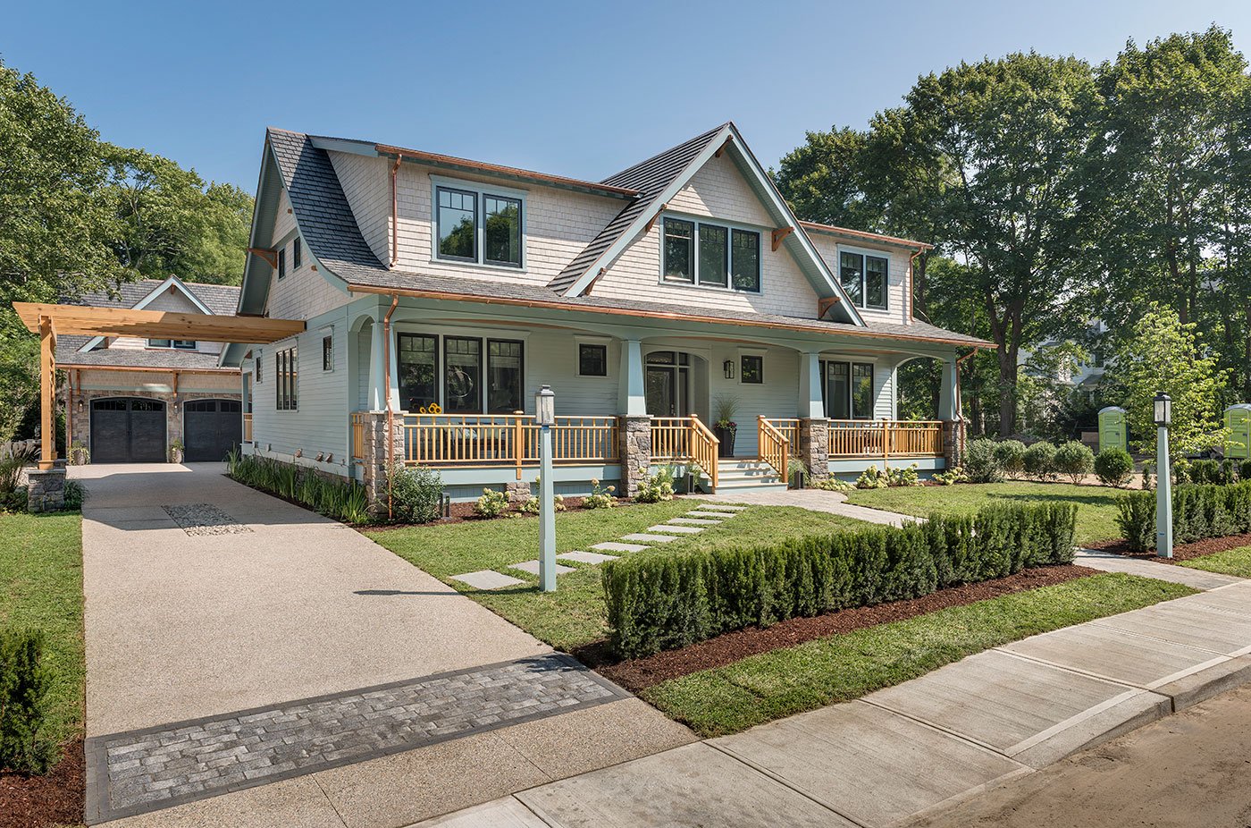 large, two-story Craftsman-style bungalow with a light-colored exterior and a prominent front porch. The house features a steep gabled roof with shingle accents and large windows that add to its charm. The porch has a wooden railing and steps leading down to a well-manicured lawn and a neatly trimmed hedge that lines the walkway. A wide driveway leads to a detached two-car garage with a matching roof. The home is surrounded by mature trees, giving it a cozy and inviting feel in a quiet suburban neighborhood.