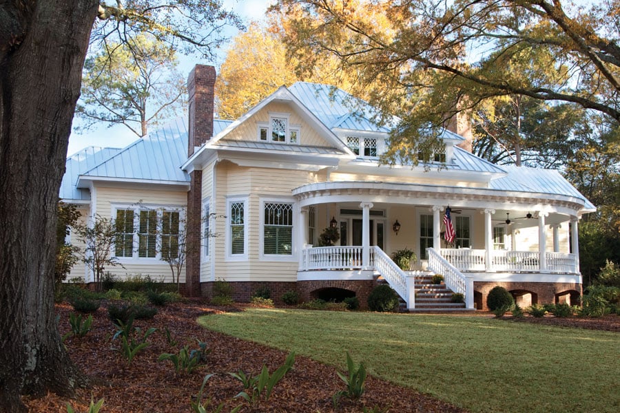 A yellow Victorian home sitting on a large lawn with a traditional, southern front porch. The home has white trim around the porch and all white windows. The windows feature diamond grille patterns.