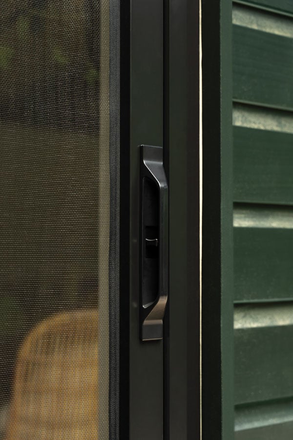 An exterior shot of a woman opening a black 200 Series Narroline® Gliding Patio Door and retractable insect screen to her deck where her dog awaits by a potted plant.