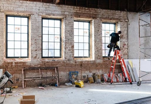 A window installer on a ladder putting the finishing touches on the fourth window in a brick-walled interior.