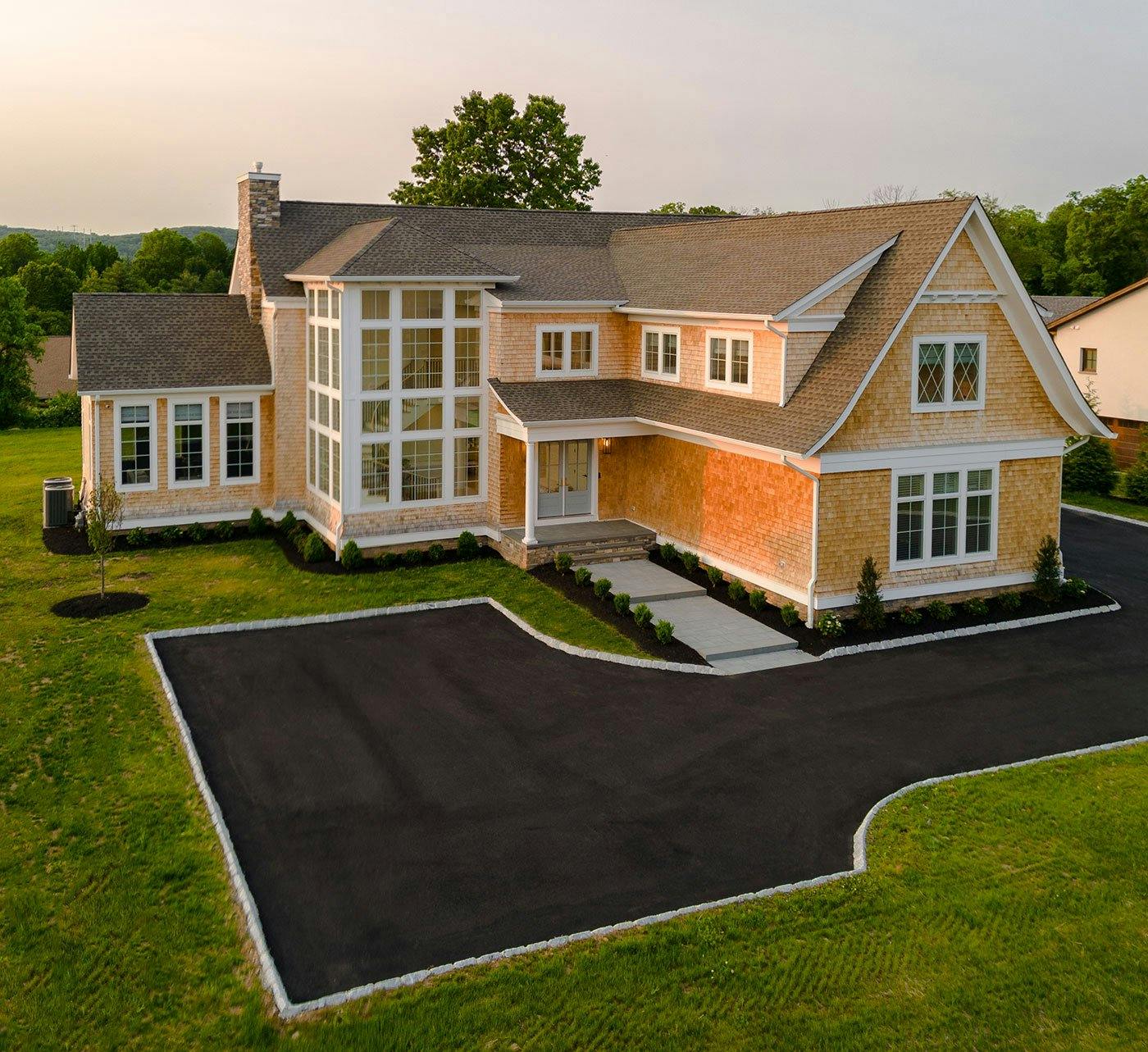 A home with brown wood shingles sits on a large plot of grassy land with a semi-circle driveway. The home has French doors and two stories of white windows with diamond and square window patterns