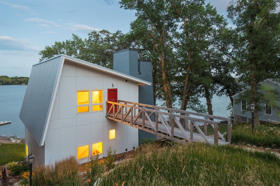 A modern lake home with white siding and metal shed roof has a second-floor entrance accessible via a wooden bridge.