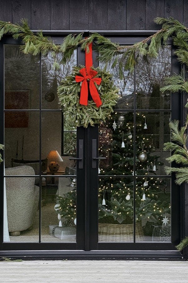 Two exterior shots of a black barn-inspired home addition with four sets of black E-Series Hinged Patio Doors that have been decorated for Christmas with evergreen boughs, wreaths, and red bows.