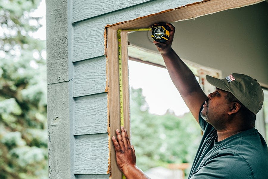 A man measures a rough opening inside a home with green wood siding. 