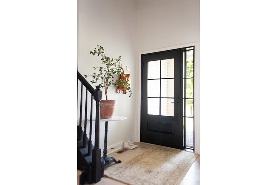  An interior shot of an entryway with a rug, table with a potted plant, and black front door whose window has colonial grilles and a sidelight to match.