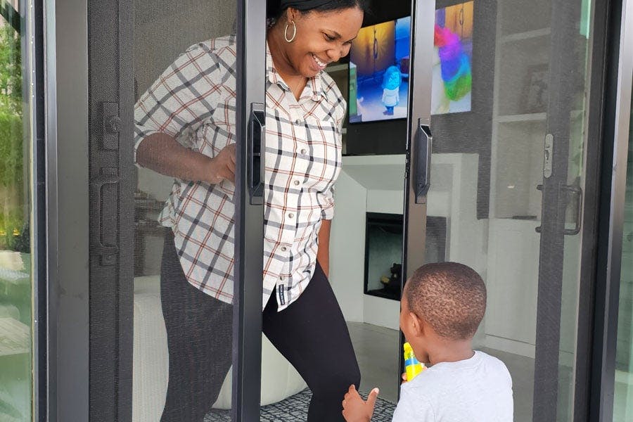 A woman is standing in the doorway of her patio door opening a retractable screen. Her two child walking up with excitement. 